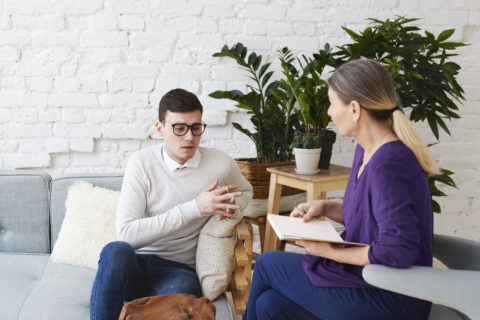 candid-shot-young-man-glasses-talking-about-his-problems-psychological-therapy-session-sitting-coach-while-mature-female-psychologist-with-copybook-listening-him-making-notes-480x320.jpg